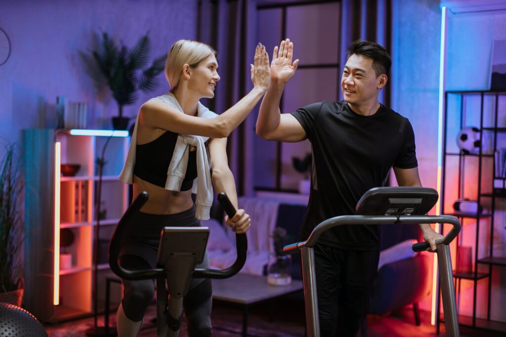 Woman training on exercise bike with asian man running on treadmill and giving high five